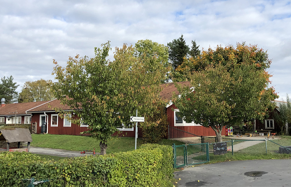 En röd träbyggnad med tegeltak. Gräsmattor och träd och en grönskande häck står framför förskolan.