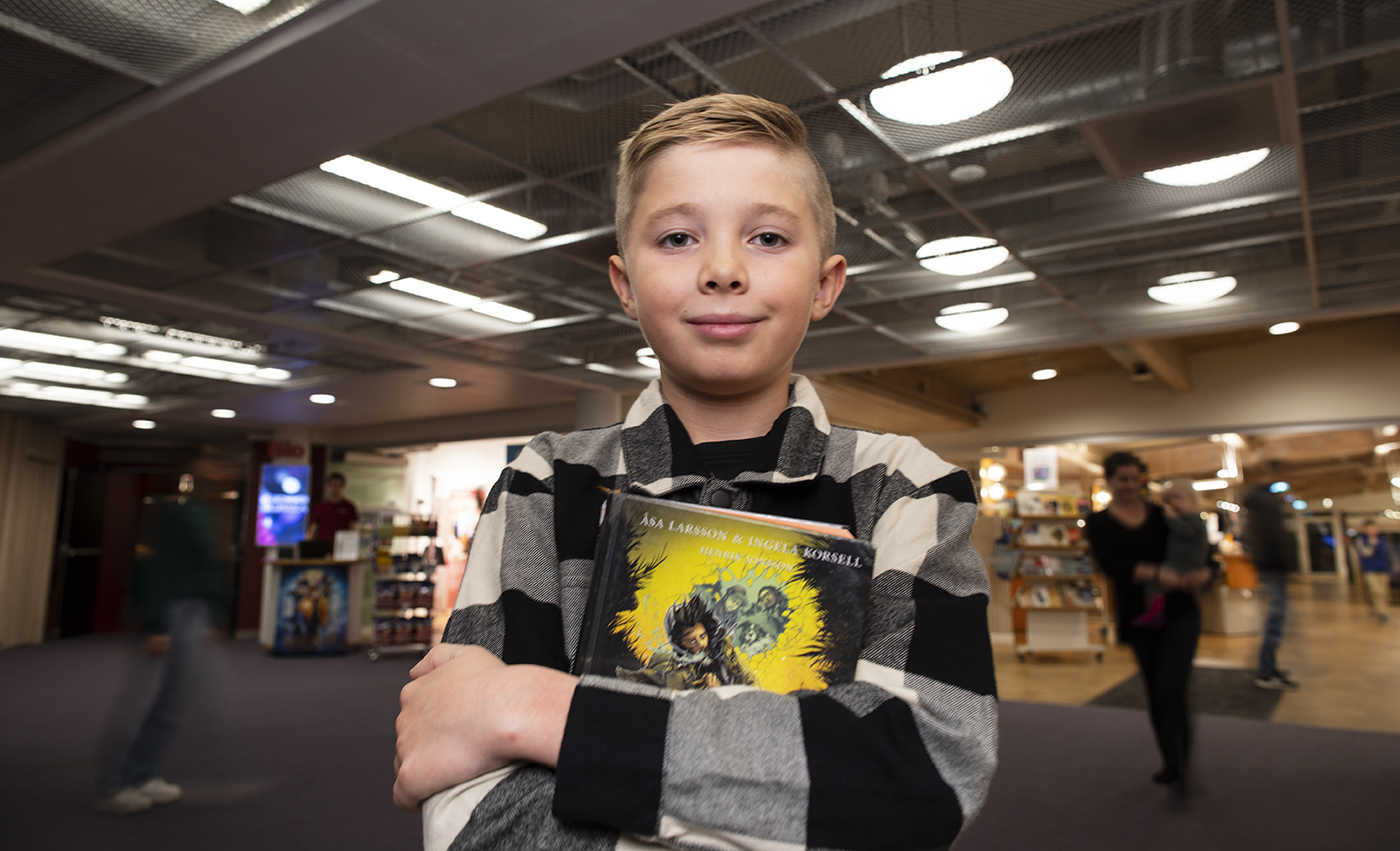 En pojke står framför biblioteket i kulturhuset och håller upp en bok.