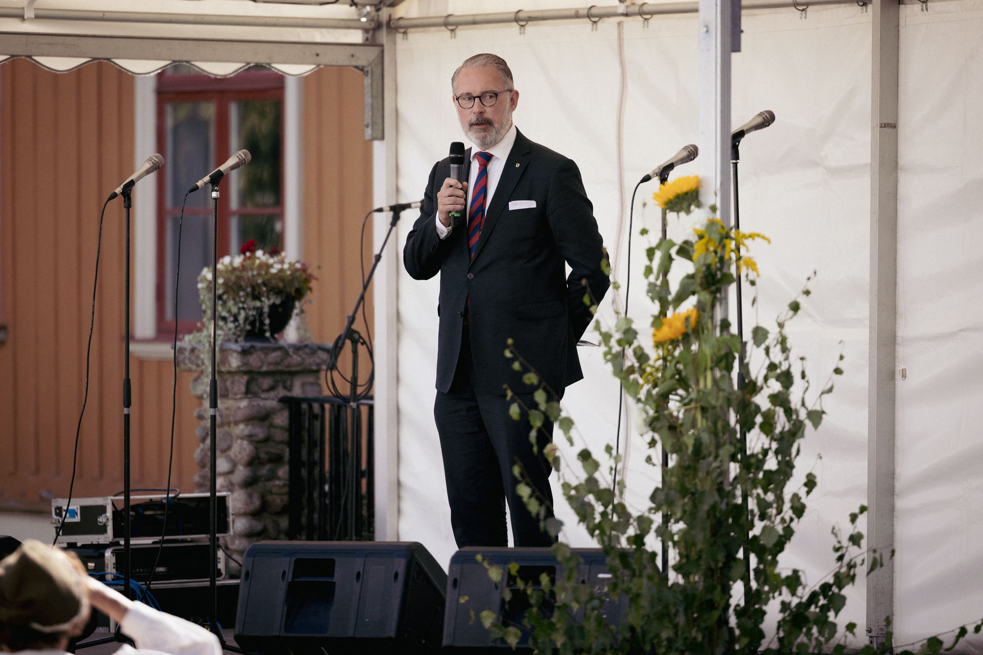 Johan Elfver (M), kommunstyrelsens ordförande står på scenen och håller tal. På scenen syns blommor.