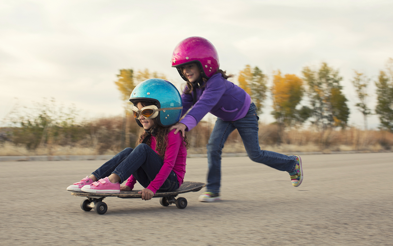 En flicka med hjälm på huvudet sitter på en skateboard. En annan flicka som också har hjälm puttar på henne med en väldig fart. Det ser ut som att de har kul.