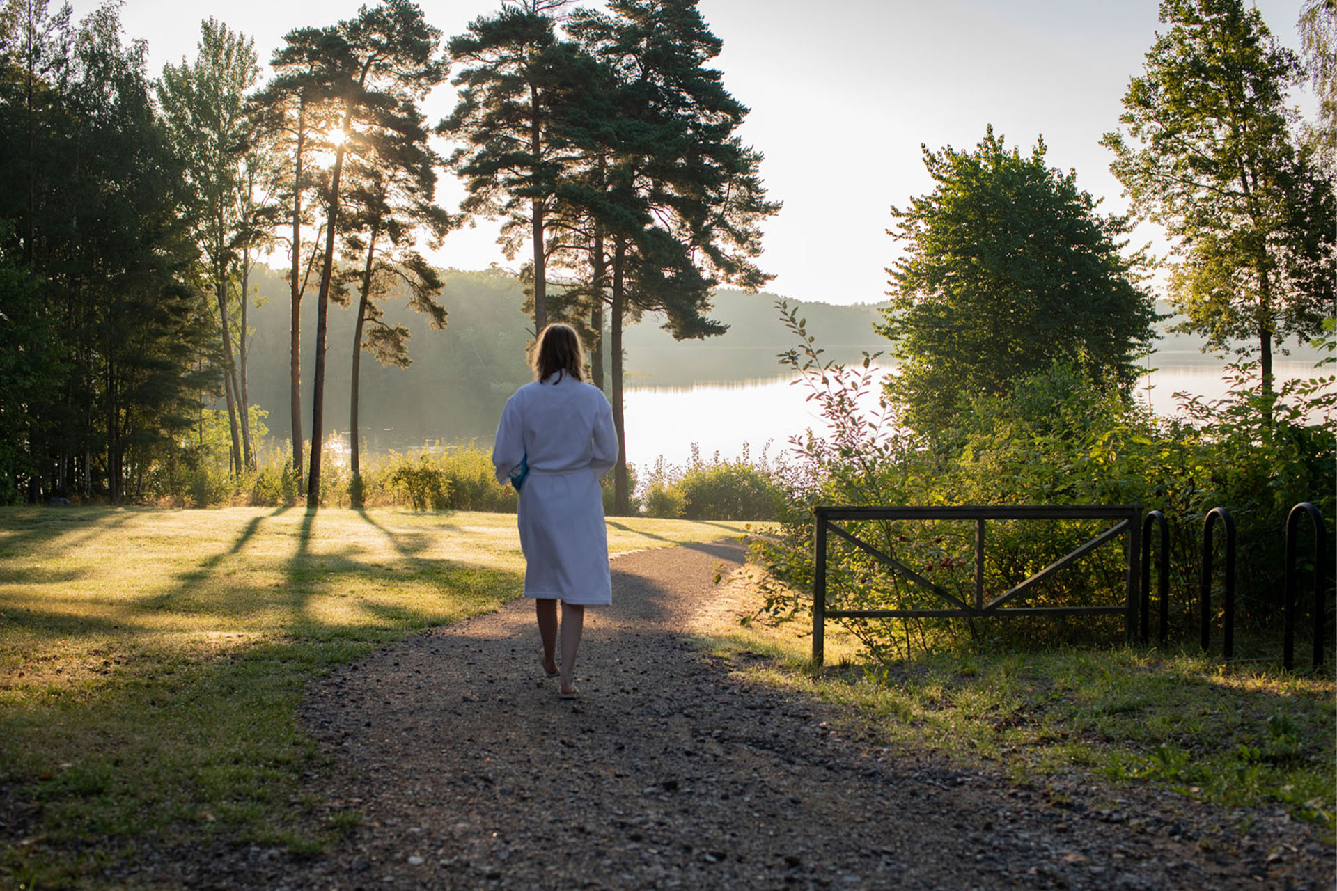 Kvinna som går på grusväg mot badplats. Klädd i vit morgonrock med buskar vid sidan och träd framför sig.
