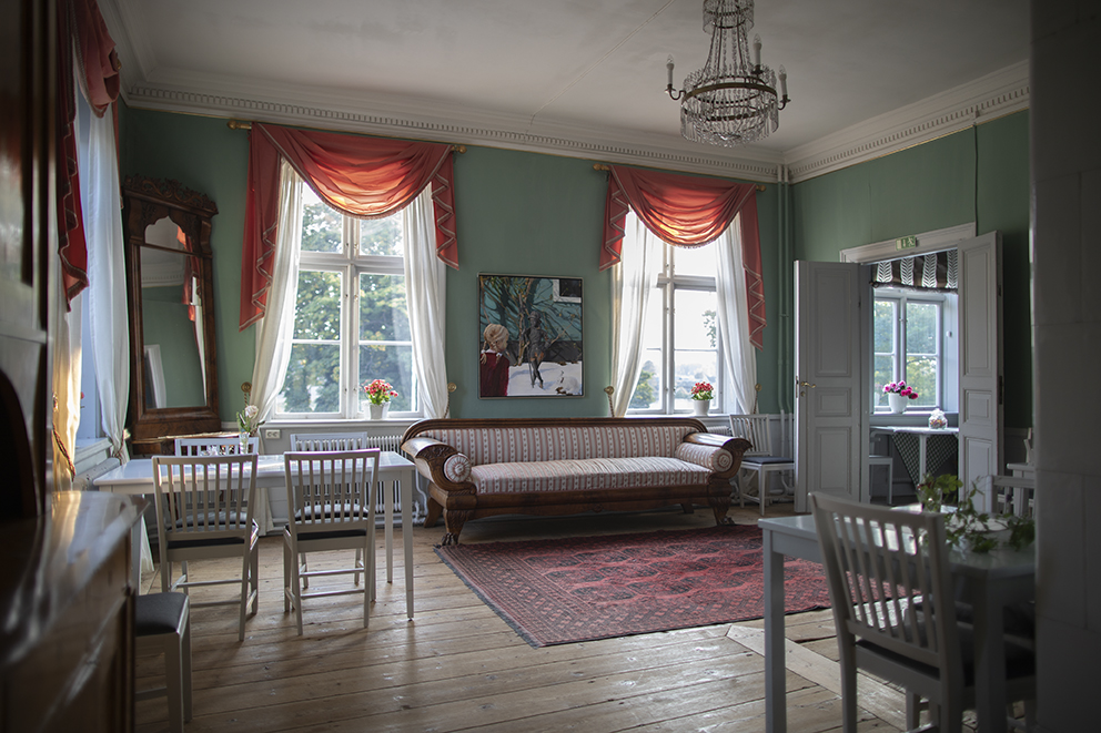 Ett grönt rum med bord och stolar. Röda gardiner hänger vid fönstren och en stor tavla hänger mellan fönstrena