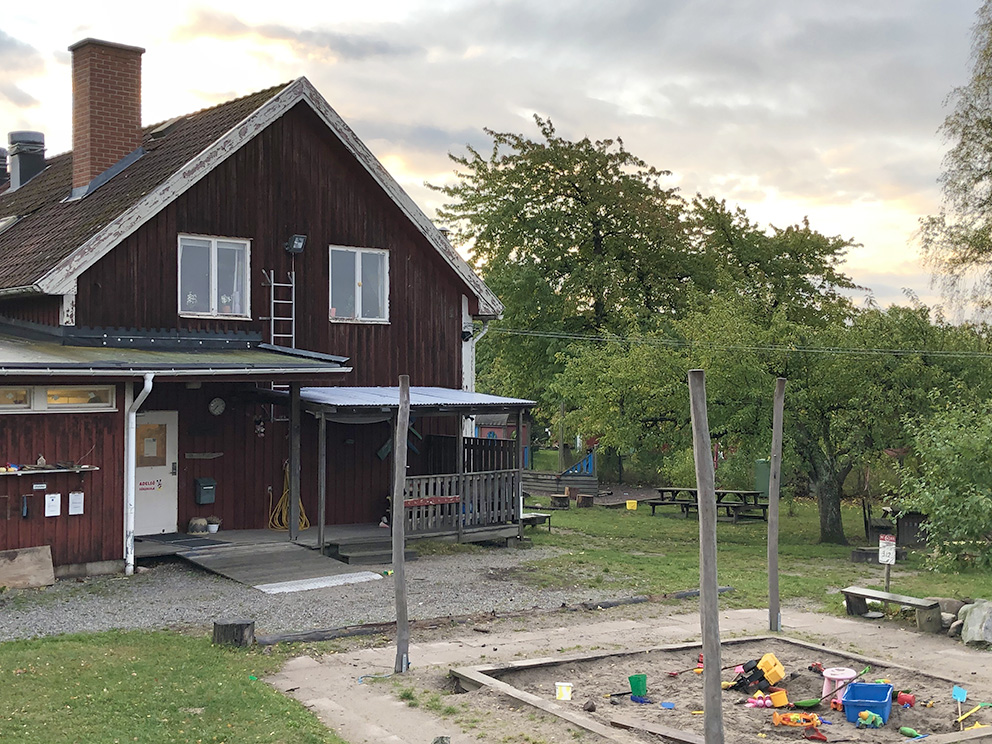 Adelsö förskola i Ekerö kommun. En röd villa med spröjsade fönster. En sandlåda med leksaker, gräs och träd.