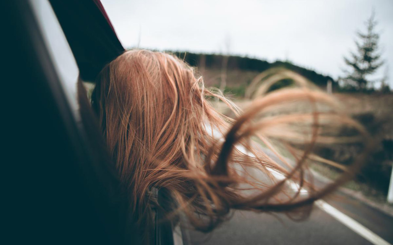 En person med långt, rött hår lutar huvudet ut genom ett bilfönster. Håret fladdrar i vinden.