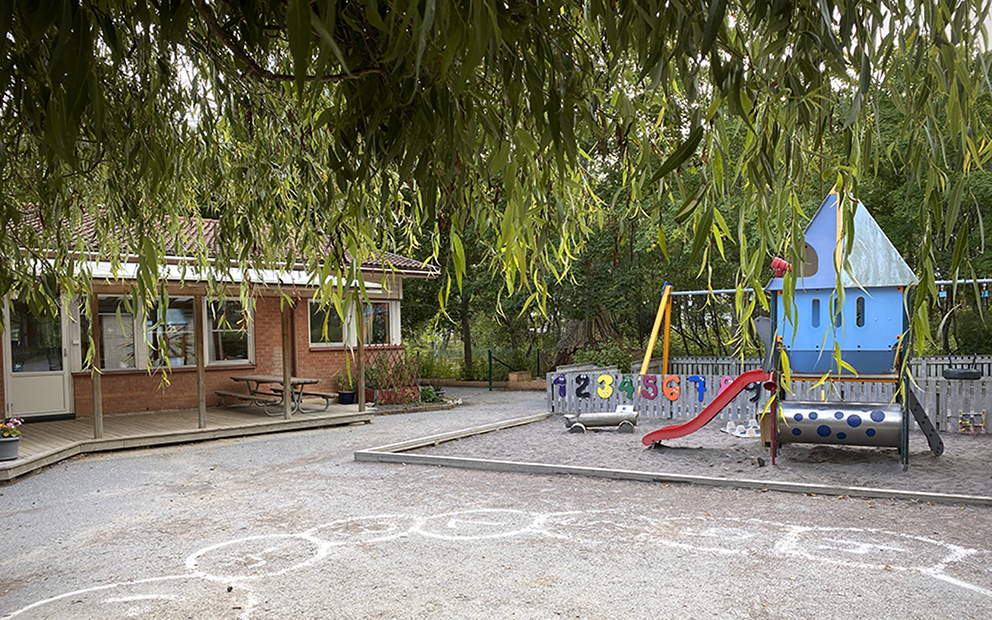 En förskolebyggnad syns i bakgrunden. En klätterställning med blått tak och en rutschkana står i en sandlåda. Man anar gungor bakom klätterställningen.