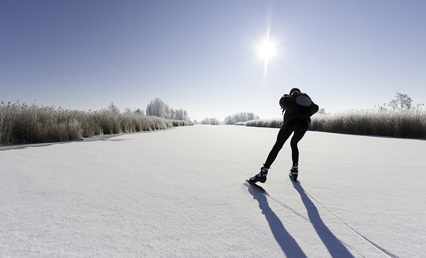 En skridskoåkare åker skridskor på en frusen sjö. Himlen är ljusblå och solen skiner. På isen ligger det snö.