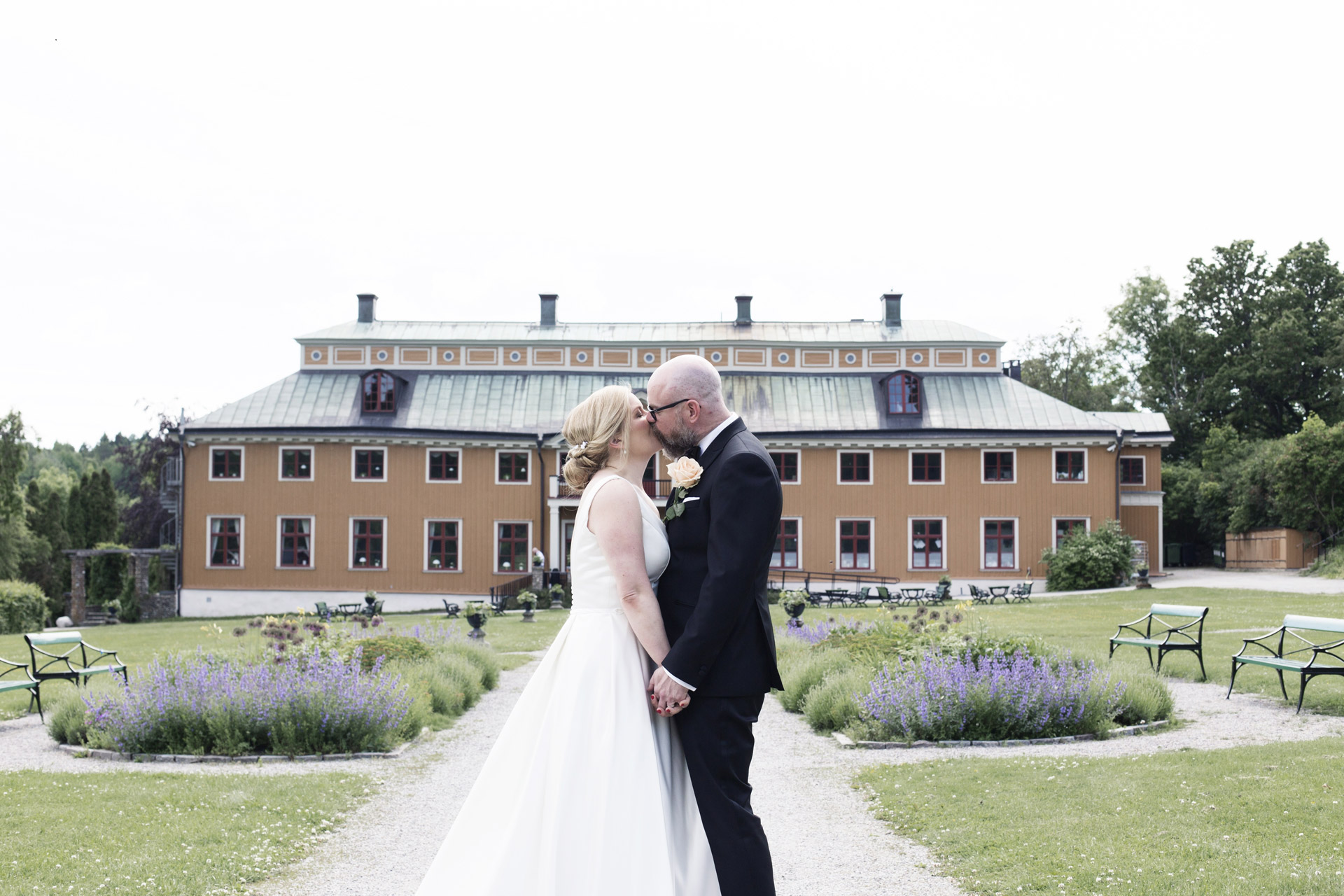 Ett bröllopspar framför Ekebyhovs slott. Det är somrigt och blålila blommor i trädgården.