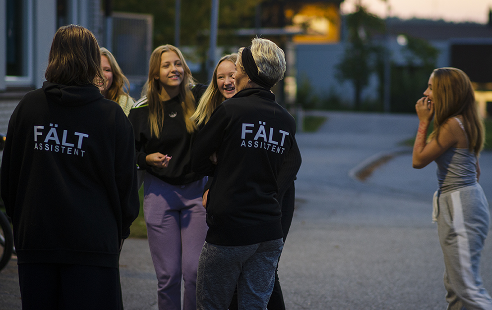 Två fältassistenter med tröjor på där det står FÄLT står och pratar med fyra unga tjejer.