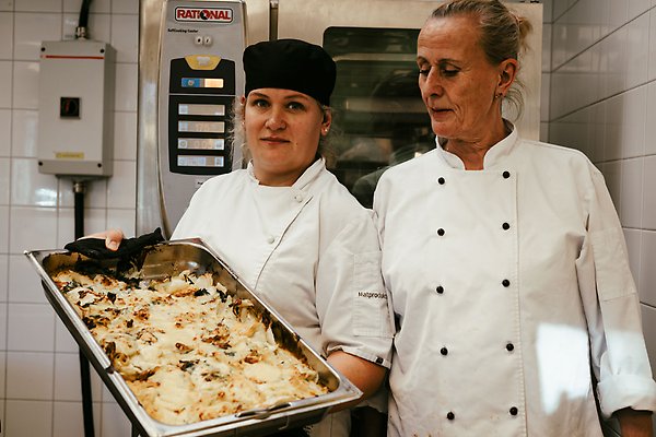 Angelica och Åse bredvid varandra i köket i Närlundaskolan. Angelica håller upp ett bläck med pastagratäng. Ugn i bakgrunden. 